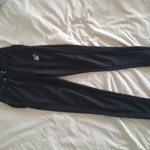 Svarta mjukisbyxor från SikSilk - Svarta mjukisbyxor från SikSilk med vit broderad logga på låret. Byxorna har resår och snörning i midjan samt smal passform med muddar vid bensluten. Perfekta för chill eller träning.