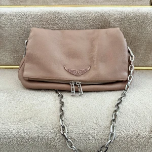 Ljusrosa axelväska från Zadig & Voltaire - Säljer en beige axelväska i mjukt skinn från Zadig & Voltaire. Väskan har en silverfärgad kedja som axelrem och dubbla dragkedjor framtill. Baksidan har ytterfack med dragkedja. Nypris 4650kr.