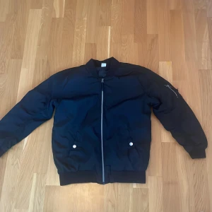 Svart bomberjacka från Lindex Kids - Svart bomberjacka från Lindex Kids i storlek 152. Jackan har klassisk bombermodell med ribbade muddar, dragkedja framtill och två fickor med tryckknappar. Materialet är syntet och jackan har en ärmficka med dragkedja för extra cool detalj.