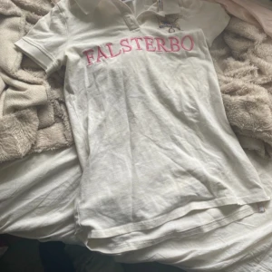 Vit piké Falsterbo x Gina Tricot XXS - Vit pikétröja från Gina Tricot i samarbete med Falsterbo Horse Show. Klassisk krage, knäppning framtill och broderad logga på bröstet. Stor rosa text 'FALSTERBO' över bröstet. Perfekt för dig som gillar häst och ridning.