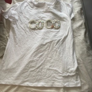 Vit t-shirt med paljetttext från Impresion - Vit t-shirt från Impresion med korta ärmar och rund hals. Framsidan har texten 'COCO' i stora bokstäver dekorerade med paljetter, pärlor och små blommor. Materialet är mjuk bomull och passformen är normal. Perfekt för dig som gillar detaljer och lite extra bling.