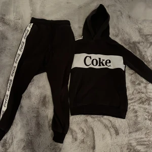 Svart Coke set hoodie med huva och byxor - Svart hoodie från Coca-Cola med stor vit panel och texten 'Coke' framtill. Tröjan har huva och en klassisk känguruficka. Perfekt för dig som gillar streetwear och vill sticka ut med en ikonisk logga.