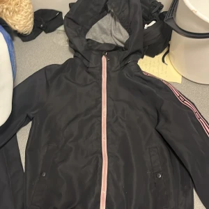 Svart vindjacka från Warp med huva - Svart vindjacka från Warp med rosa dragkedja och rosa detaljer längs ärmarna. Jackan har huva, fickor med tryckknapp och sportig look. Perfekt för dig som gillar streetstyle och vill ha något snyggt och funktionellt.