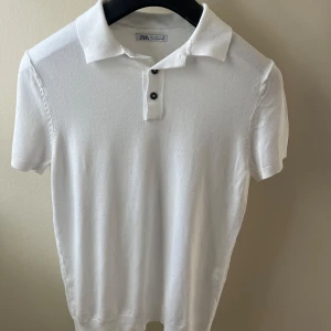 Vit pikétröja från Zara - Klassisk vit pikétröja från Zara med korta ärmar och krage. Tröjan har två svarta knappar framtill och är tillverkad i mjuk bomull. Perfekt för en clean och stilren look, passar lika bra till jeans som till shorts.