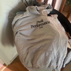 Ljusgrå hoodie från Peak Performance - Säljer en ljusgrå hoodie från Peak Performance med svart logga på bröstet. Tröjan har huva, magficka och ribbade muddar. Perfekt för chill dagar eller när du vill ha en clean och enkel stil.