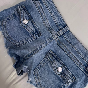 Blå jeansshorts med råa kanter - Snygga blå jeansshorts från Gina tricot med råa, fransiga kanter och coola fickdetaljer bak med knappar. Klassisk femficksmodell och bälteshällor. Har haft hål bak men som blivit ihop sydda. Perfekta för varma dagar. 