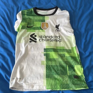 Liverpool bortatröja Szoboszlai #8 Nike - Liverpool FC bortatröja med Szoboszlai #8 på ryggen. Tröjan är vit med gröna grafiska mönster och svarta detaljer. Nike-logga, klubbmärke och sponsortryck på bröstet. Tillverkad i lätt och ventilerande material, perfekt för match eller träning.