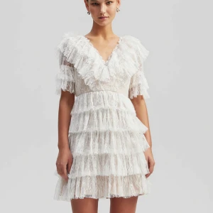 Vit spetsklänning med volanger - Superfin kort klänning i vit spets med flera lager volanger och v-ringning. Klänningen har korta ärmar och romantisk känsla, perfekt för dig som gillar en söt och feminin stil. Materialet är spets och den har en figurnära passform.