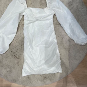 Vit kort klänning från Nelly  - Supersnygg vit kort klänning från Nelly med puffiga långärmar och figurnära passform. Klänningen har fyrkantig ringning och är gjord i ett lätt, slätt material som ger en fräsch look. Perfekt för dig som vill sticka ut med stilrena detaljer.