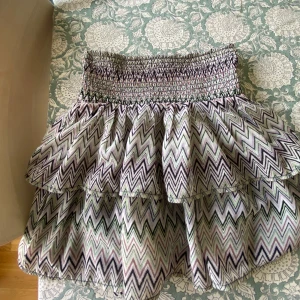 Kort volangkjol med zigzag-mönster - Supersöt kort kjol med volanger och elastisk smock i midjan. Kjolen har ett färgglatt zigzag-mönster i svart, lila, grön och vit. Perfekt för dig som gillar en lekfull och trendig stil. Lätt och luftig, passar perfekt till sommaren.