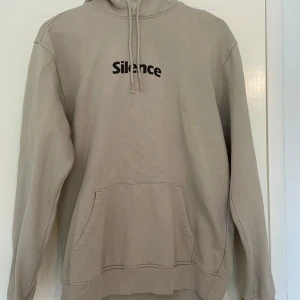 Beige hoodie Silence med huva - Säljer en stilren beige hoodie från Silence med svart tryck på bröstet. Tröjan är använd fåtal gånger och har tyvärr några fläckar men de syns knappt. 
