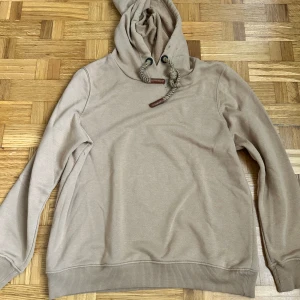 Beige hoodie från COOD med grova snören - Beige hoodie från COOD med stora grova snören i huvan och snygga läderdetaljer. Tröjan har ribbade muddar och en avslappnad passform. Perfekt för chill dagar och enkel att matcha med jeans eller joggers.