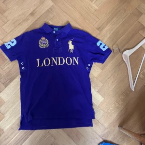 Lila pikétröja från Polo Ralph Lauren - Säljer en lila pikétröja från Polo Ralph Lauren med broderad logga och texten LONDON i gult framtill. Tröjan har korta ärmar, krage och knappar vid halsen. Perfekt för en sportig och stilren look. Den är legit checkad då de flesta inte är äkta som säljs
