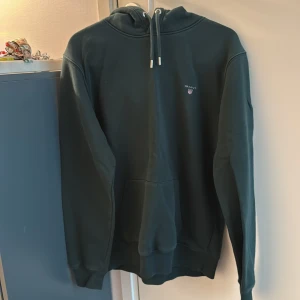 Mörkgrön hoodie från GANT - Säljer en mörkgrön hoodie från GANT i storlek L. Tröjan har huva med dragsko, magficka och diskret GANT-logga på bröstet. Perfekt för en avslappnad stil och enkel att matcha med det mesta.