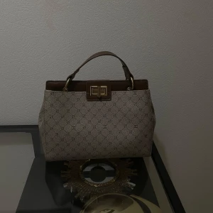Beige och brun klassisk handväska  - Säljer en snygg beige och brun handväska med klassiskt monogrammönster och guldfärgat spänne. Väskan har ett stadigt handtag och detaljer i brunt skinnliknande material. Perfekt accessoar för att lyfta din outfit. Har tyvärr för många väskor hemma 