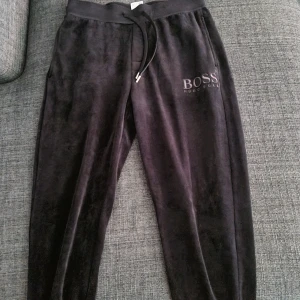 Svarta mjukisbyxor från Hugo Boss - Svarta mjukisbyxor från Hugo Boss med dragsko i midjan och logga tryckt på ena benet. Byxorna har en mjuk och skön känsla, perfekta för avslappnade dagar. Klassisk passform och elastisk midja.