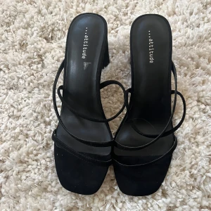 Svarta klackar med smala remmar från Attitude - Säljer ett par svarta klackar från Attitude med smala remmar och fyrkantig tå. Skorna har öppen tå och hög klack, perfekta för att lyfta din outfit. Snygg minimalistisk design med trendig siluett.