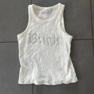 Vit ribbad linnetopp med strass från Lindex - Säljer en vit ribbad linnetopp från Lindex med texten 'Bright' i strass framtill. Toppen är ärmlös och har en klassisk rund halsringning. Perfekt för varma dagar eller som lager under andra plagg.