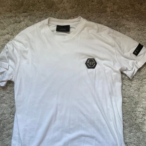 Vit t-shirt från Philipp Plein - Snygg vit t-shirt från Philipp Plein med svart PP-logga på bröstet och metallbokstäver på ärmen. Klassisk rund hals och normal passform. Perfekt för dig som gillar exklusiva streetwear-plagg.