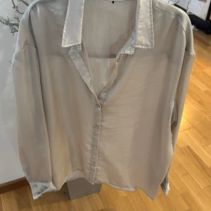 Beige satinblus från Lefties - Säljer en stilren beige blus i satin från Lefties. Blusen har klassisk krage, knäppning framtill och långa ärmar med knapp vid ärmslut. Perfekt för dig som gillar en elegant och enkel look.