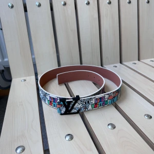 Louis Vuitton bälte med färgglatt mönster - Säljer ett Louis Vuitton bälte med svart spänne och färgglatt, tecknat mönster med LV-logga. Bältet är gjort i skinn och har en vit bas med inslag av blått, rött och svart. Märkt 'Louis Vuitton Paris made in France' på insidan.