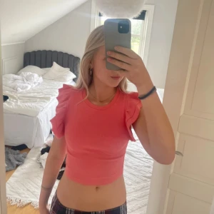 Korallrosa croppad topp med volangärmar - Säljer en söt rosa croppad topp med volangdetaljer på ärmarna. Toppen har en tight passform och är perfekt för sommaren. Superfin att styla till både jeans och kjol!