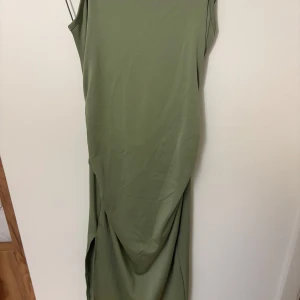 Olivgrön långklänning med rosettdetalj från SHEIN - Säljer en olivgrön långklänning från SHEIN med smala axelband och snygg rosettdetalj framtill. Klänningen har slits på sidan och är figurnära med en elegant siluett. Perfekt för dig som vill sticka ut med en stilren look.