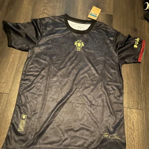 Svart Nike fotbollströja med guldiga detaljer - Säljer en svart fotbollströja från Nike med guldiga detaljer och tryck. Tröjan har korta ärmar, rund hals och diskreta mönster i tyget. Märket syns på bröstet och det finns även små röda detaljer på ärmsluten.