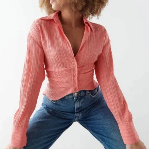 Korrallrosa skjorta med knappar - Säljer en snygg korrallrosa skjorta med lång ärm och markerad midja. Skjortan har knappar framtill och är tillverkad i ett lätt, strukturerat tyg som ger en cool look. Perfekt att styla med jeans eller kjol.