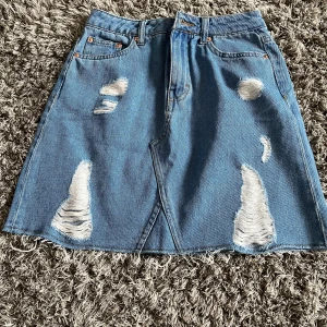 Blå jeanskjol fråns Divided - Säljer en blå jeanskjol från Divided med slitna detaljer framtill och klassiska fickor både fram och bak. Kjolen har en A-linjeform och är kort i modellen. Perfekt för dig som gillar en avslappnad och trendig stil.