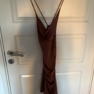 Brun långklänning med smala axelband - Säljer en snygg brun långklänning med smala axelband och djup v-ringning. Klänningen har draperad effekt framtill och en figurnära passform. Köpt på Bikbok💕