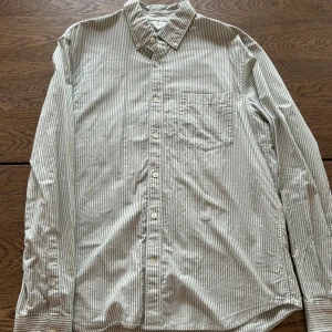 Grönvit randig skjorta från Arket - Snygg skjorta i grön och vitt med smala ränder från Arket. Klassisk krage, bröstficka och knäppning framtill. Skjortan är långärmad och tillverkad i mjuk bomull. Storlek EU 50, vilket passar bra för de som normalt sett har storlek M/L. Mycket fint skick.