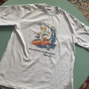 Vit t-shirt med surfande björn från Polo Ralph Lauren - Vit t-shirt från Polo Ralph Lauren! As nice för sommaren och aldrig använd! Prima skick och endast lite skrynklig! Passar S men också M! Skriv för frågor🙌
