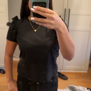 Svart t-shirt med volang från Zara - Säljer en svart t-shirt från Zara med volangdetaljer på axlarna. T-shirten har rund halsringning och korta ärmar. Perfekt att styla till jeans eller kjol för en snygg look.