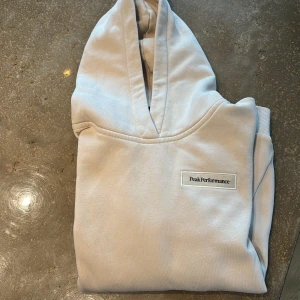 Beige hoodie från Peak Performance - Säljer en stilren beige hoodie från Peak Performance med broderad logotyp på bröstet. Tröjan har huva, långa ärmar och en klassisk känguruficka framtill. Perfekt för en avslappnad och trendig look.