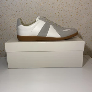 Maison Margiela gats  - Säljer ett par Maison Margiela gats sneakers i vitt skinn med grå mockadetaljer och beige sula. Klassisk design med snörning och rund tå. Perfekta för dig som gillar stilrena och tidlösa sneakers.