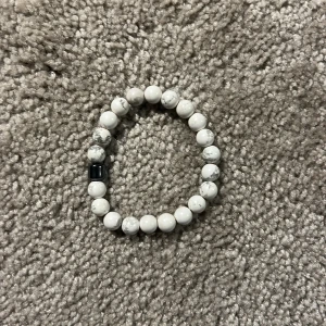 Vitt pärlarmband med svart detalj - Stilrent armband med vita pärlor och en svart pärldetalj. Elastiskt band som gör det enkelt att ta på och av. Perfekt accessoar för att lyfta din outfit med en enkel men snygg touch.