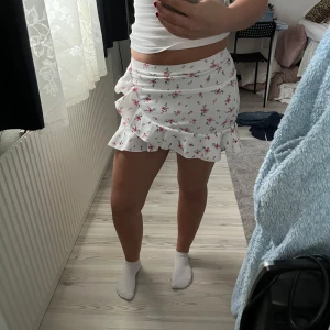 Vit blommig omlottkjol med volang - Supersöt vit omlottkjol med rosa blommor och volangkant. Kjolen har en feminin och somrig känsla, perfekt för varma dagar. Den är kort och har en omlottmodell