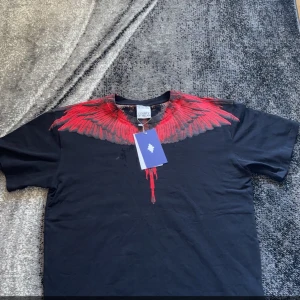 Svart t-shirt Marcelo Burlon - Svart t-shirt från Marcelo Burlon med ett iögonfallande rött vingmönster över axlar och hals. T-shirten har normal passform och är kortärmad. Perfekt för dig som vill sticka ut med en unik design.