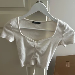 Vit croppad topp från Zara - Säljer en vit croppad topp från Zara med korta ärmar och ribbad struktur. Toppen har en snygg v-ringning och knytning framtill som ger en trendig look. Perfekt att matcha med jeans eller kjol!