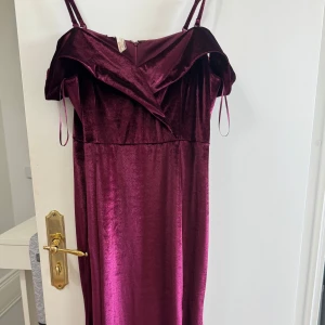 Vinröd sammet långklänning med offshoulder - Vinröd långklänning i mjuk sammet med smala axelband och offshoulder-detalj. Klänningen har en elegant v-ringning och en figurnära passform upptill. Perfekt för festliga tillfällen. Oanvänd