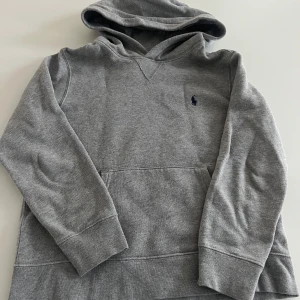 Grå hoodie från Polo Ralph Lauren 10-12 år - Säljer en klassisk grå hoodie från Polo Ralph Lauren med broderad logga på bröstet. Tröjan har huva, lång ärm och en stor magficka framtill. Perfekt för en avslappnad och stilren look.