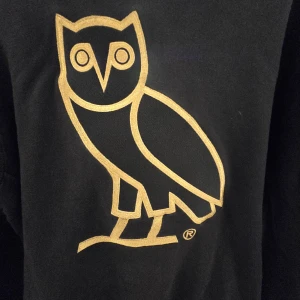 Svart hoodie från OVO med guldfärgad uggla stl L - Säljer en svart hoodie från OVO med deras ikoniska ugglelogo i guld på bröstet. Tröjan har en klassisk passform och är perfekt för dig som gillar streetwear och snygga detaljer. Stl L