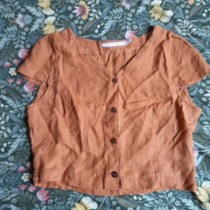 Brun kortärmad blus från Pennyblack - Säljer en snygg brun kortärmad blus från Pennyblack i 100% linne. Blusen har v-ringning och knappar framtill, perfekt för varma dagar. Enkel och lite vintage design.