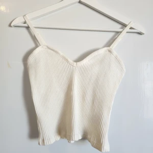 Vit ribbad linnetopp från Zara - Säljer en vit ribbad linnetopp från Zara med smala axelband och lätt v-ringad halsringning. Toppen är croppad och har en figurnära passform, perfekt till sommaren. Justerbara axelband.