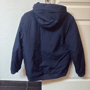 Mörkblå pufferjacket från Nike med huva - Säljer en mörkblå pufferjacket från Nike med huva och dragkedja framtill. Jackan har två sidofickor och en liten logga på bröstet. Perfekt för blåsiga dagar och enkel att matcha med det mesta. Har tre väldigt små hål på framsidan-syns på bild 4-5