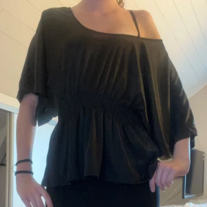 Svart offshoulder topp - Svart topp med lös passform och offshoulder-design. Ena axeln är bar och den har ett brett resårband i midjan som ger en snygg siluett. Perfekt för dig som gillar en avslappnad men trendig stil.