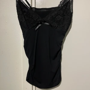 Svart spetslinne med rosett - Snyggt svart linne med spets upptill och en liten rosett framtill. Axelbandslös design med feminina detaljer och mjukt material. Perfekt att styla till olika outfits.