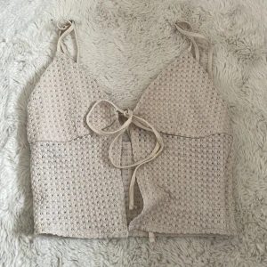 Beige croppad linnetopp från H&M i xxs - Säljer en beige croppad linnetopp från H&M med smala axelband och knytning framtill. Toppen har ett diskret hålmönster i tyget och justerbara axelband för perfekt passform. Tänk på att den är väldigt kort så man kan ha den över ett linne eller bikini! Säljer då den aldrig kommit till användning.  