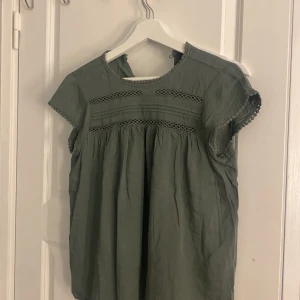Grön blus med spetsdetaljer - Säljer en söt grön blus med spetsdetaljer vid ärmar och bröst. Blusen har korta ärmar och en lös passform, perfekt för vår och sommar. Tillverkad i mjukt bomullsmaterial.